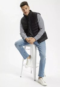 Czarna kamizelka puchowa na szarej koszuli w kratę, w zestawie z jasnoniebieskimi dżinsami i białymi sneakerami, siedzący na białym stołku.