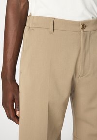 Beige maßgeschneiderte Hosen aus glattem Stoff, mit einem klassischen Knopfverschluss und einem eleganten Design, mit sichtbaren Seitentaschen.