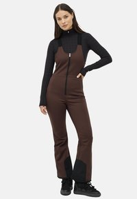 Brun skiftedragt med lynlås foran, justerbare stropper og sorte detaljer ved anklerne. Båret over en tætsiddende sort langærmet bluse.