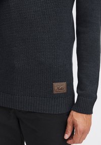 Donker, gebreide trui met een gestructureerde weving, geribbelde manchetten en een bruine leren label aan de onderrand. De kleur is diep marineblauw.