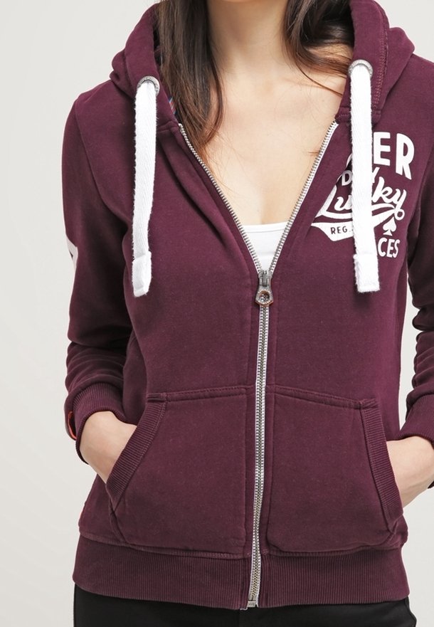 Sudadera con capucha color burdeos con cremallera, cordones blancos, bolsillos delanteros, puños acanalados y detalle de logo en el pecho izquierdo. Textura de tejido suave.