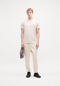 Beige póló kis fekete logóval, világos bézs melegítőnadrággal párosítva. A modell egy szürke táskát tart, fekete sportcipőt visel. Semleges háttér.