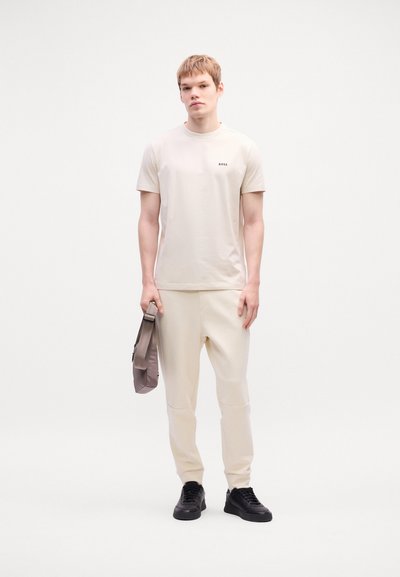T-shirt beige avec un petit logo noir, associé à un jogging beige clair. Le modèle tient un sac gris et porte des baskets noires. Fond neutre.