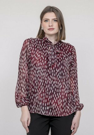 Femme aux cheveux mi-longs portant des boucles d'oreilles argentées et un chemisier à manches longues bordeaux avec un motif géométrique et un nœud au col, associé à un pantalon noir.