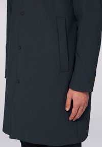 Tmavě šedý nepromokavý kabát s elegantním designem, vybavený knoflíkovým zapínáním, boční kapsou a nenápadným akcentem s logem na manžetě.