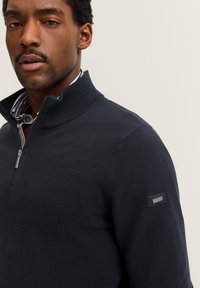 Mann in marinefarbener, strukturiertem Viertelreißverschluss-Pullover über gestreiftem Hemd mit Kragen, mit Markenlabel auf dem linken oberen Ärmel, neutraler Hintergrund.