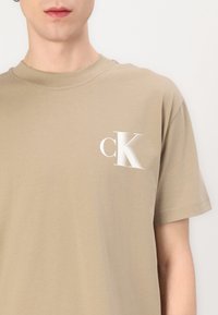 Beige medvilninė marškinėliai su apvalia iškirpte, trumpomis rankovėmis ir balta "cK" logotipu ant krūtinės. Minkšta tekstūra ir laisvas pritaikymas.