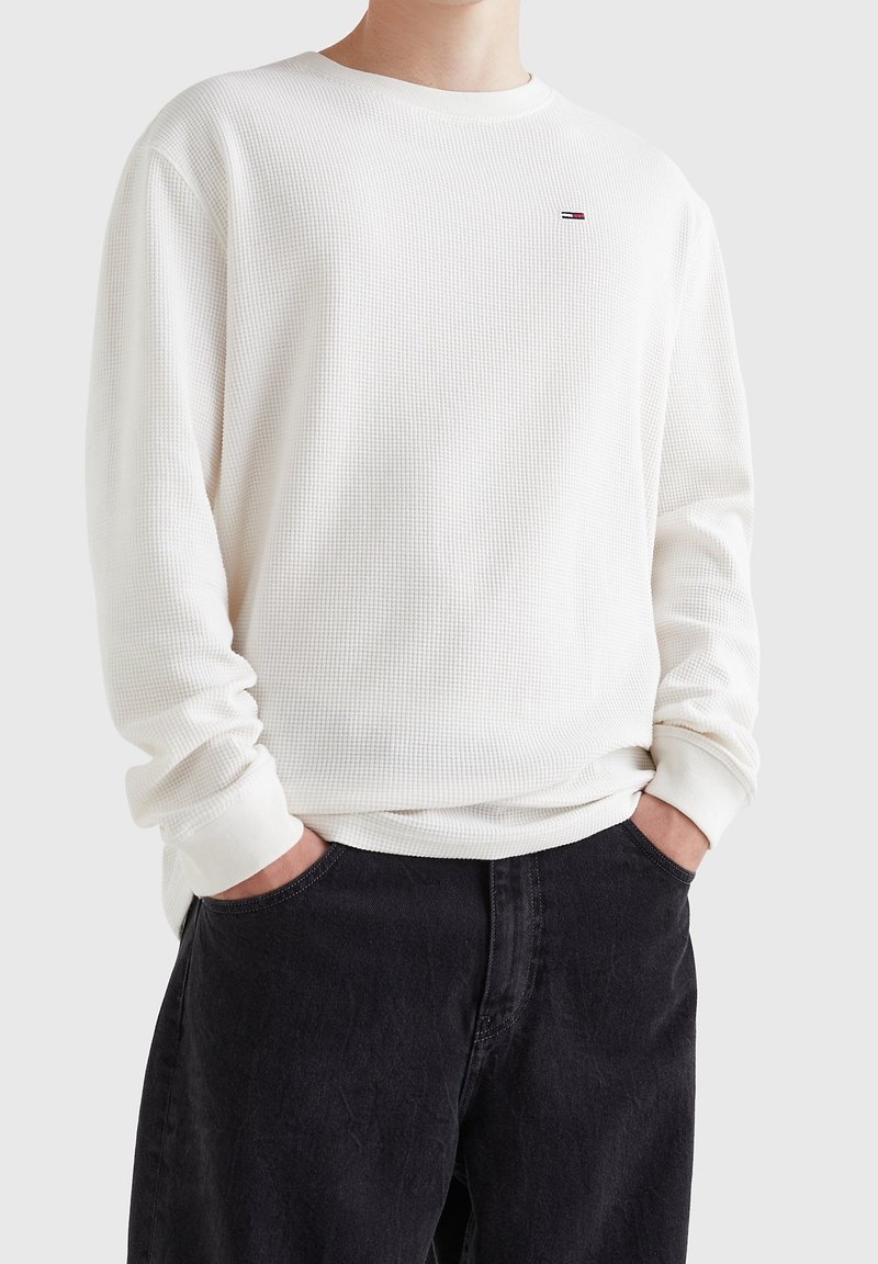 Bílé dlouhé tričko s rukávy vyrobené z texturované bavlny, s kulatým výstřihem a ribbenovými manžetami, zdobené malým logem na hrudi.