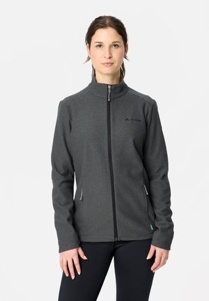 Femme portant une veste zippée texturée gris foncé, debout devant un fond clair uni.