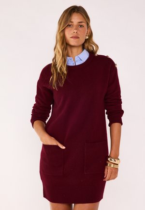 Frau trägt ein burgunderrotes Strickkleid mit Fronttaschen, goldenen Knopfdetails an der Schulter, hellblauem Hemdkragen sowie goldenen Armbändern und Ohrringen.