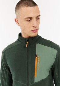 Jonge man met kort haar die een donkergroene ritsjack draagt met een lichtgroene paneel en oranje rits op de borst.