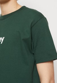 T-shirt en coton vert foncé à manches courtes et col rond, avec du texte blanc sur le devant. Texture lisse et coupe décontractée.