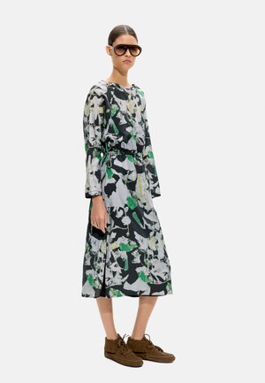 Kobieta w dużych okularach przeciwsłonecznych, długiej sukience w czarno-biało-zielony wzór z wcięciem w talii oraz brązowych mokasynach, stojąca.