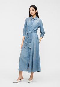 Φόρεμα πουκάμισο μεσαίου μήκους από τζιν, με ζώνη στη μέση, κουμπιά μπροστά και γιακά. Υφασμα σε ανοιχτό μπλε με διακριτική υφή.