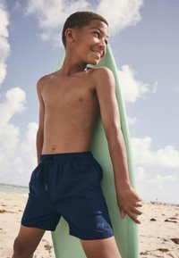 Námornícky modré plavkové šortky s elastickým pásom a sťahovacou šnúrkou, s malým logom. Chlapec stojí s zeleným surfovým prknom na piesočnatej pláži.