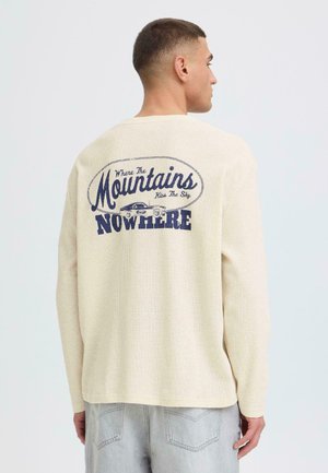 Uomo che indossa un maglione crema con testo blu navy e grafica di un'auto sul retro con la scritta "Dove le montagne baciano il cielo, OVUNQUE."