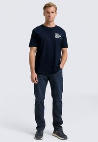 Laivastonsininen puuvillainen t-paita, jossa on lyhyet hihat ja pyöreä kaulus, vasemmalla puolella valkoinen logo, yhdistetty tummansinisiin farkkuihin ja lenkkareihin.