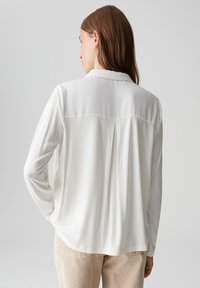 Weißes Langarmbluse mit Kragen, ausgestattet mit einer glatten Textur und einer Falten-Details an der Rückseite, kombiniert mit hellbeigen Hosen.