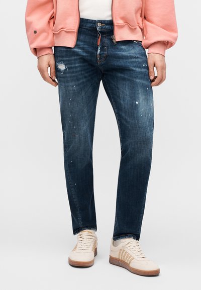 Μπλε τζιν παντελόνι με ξεθωριασμένο φινίρισμα, με φθαρμένες λεπτομέρειες και πιτσιλιές από μπογιά. Συνδυασμένο με ανοιχτόχρωμα αθλητικά παπούτσια.