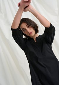 Moterys su trumpais tamsiais plaukais, dėvinčiais juodą ilgomis rankovėmis palaidinę, pakelia rankas virš galvos prieš šviesų neutralų audinio foną.