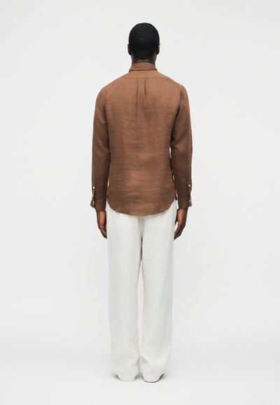 Άνδρας με την πλάτη προς τα έξω, φοράει καφέ μακρυμάνικο πουκάμισο και άνετο λευκό παντελόνι, στέκεται μπροστά σε ένα απλό ανοιχτό φόντο.