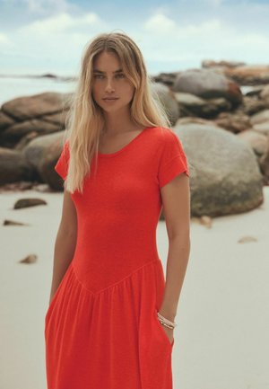 Tânără cu păr blond lung, purtând o rochie roșie cu mâneci scurte, stând pe o plajă nisipoasă cu stânci mari în fundal.