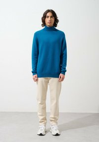 Maglione turtleneck turchese con polsini e orlo a costine, abbinato a pantaloni color crema e sneaker bianche, in piedi contro uno sfondo neutro.