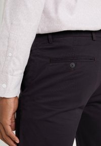 Ciemnofioletowe spodnie o gładkiej fakturze, z tylną kieszenią zapinaną na guzik. Częściowo widoczna jest jasna koszula o lekkiej fakturze.