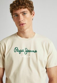 Bežinė medvilninė marškinė su žaliu "Pepe Jeans" logotipu, o žemiau baltu tekstu "LONDON", trumpomis rankovėmis ir apvaliu kaklu.