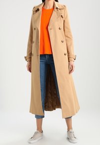 Trench coat beige con davanti doppiopetto, colletto a punte e maniche lunghe. Abbinato a un top arancione e jeans blu a vita alta.