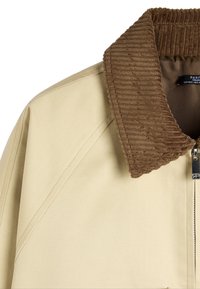 Beżowa kurtka z brązowym kołnierzem z korduroju i metalowym zamkiem z przodu, odsłaniająca ramię i górną część klatki piersiowej.