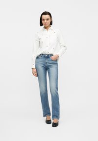 Γυναίκα που στέκεται με το ένα χέρι στην τσέπη, φοράει λευκό πουκάμισο με κόκκινα σχέδια καρδιών, μπλε τζιν και μαύρες μπαλαρίνες.