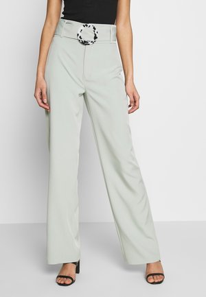 Femme portant un pantalon large gris clair avec une boucle de ceinture circulaire noire et blanche à pois et des escarpins noirs à bout ouvert.