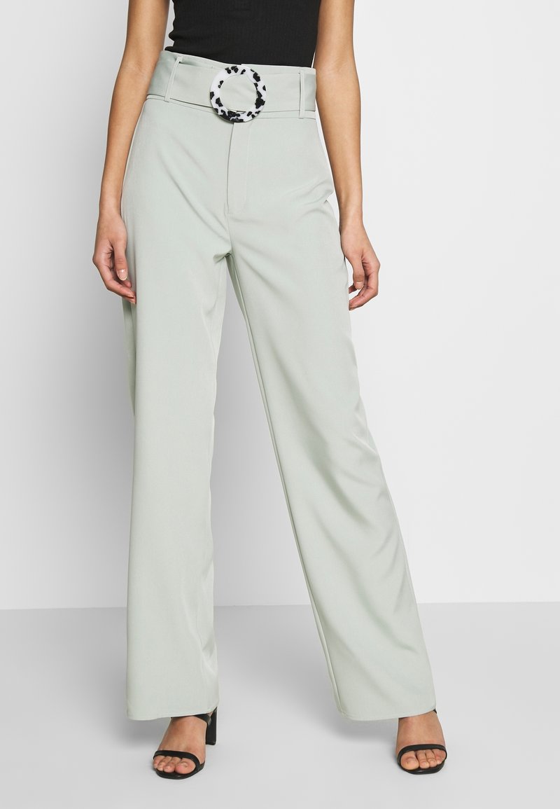 Femme portant un pantalon large gris clair avec une boucle de ceinture circulaire noire et blanche à pois et des escarpins noirs à bout ouvert.