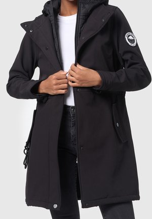 Femme portant un long manteau d'hiver noir avec capuche, en train de fermer la fermeture éclair frontale, écusson logo sur la manche gauche, chemise blanche et pantalon noir.