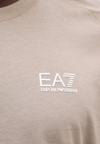 Μπεζ ύφασμα με λευκό λογότυπο EA7 Emporio Armani τυπωμένο στην περιοχή του στήθους ενός ενδύματος.