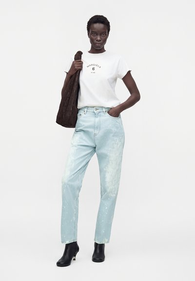Γυναίκα που φοράει λευκό t-shirt με το λογότυπο Margiela, ανοιχτόχρωμο τζιν με πιτσιλιές βαφής, μαύρα μποτάκια στον αστράγαλο, κρατώντας καφέ υφασμάτινη τσάντα ώμου.