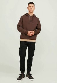 Brun pullover-hoodie med en logga framtill, beige lager under, kombinerad med svarta cargobyxor och bruna sneakers med vita sulor.