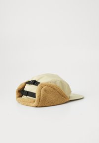 Beige hat with a soft, tan fleece lining and ear flaps. Features adjustable black fabric straps for secure fit and a curved visor.