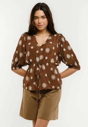 Kobieta o długich ciemnych włosach, ubrana w brązową bluzkę w kwiaty z bufkami i wiązaniem w talii, do tego krótkie spodenki khaki sięgające kolan.