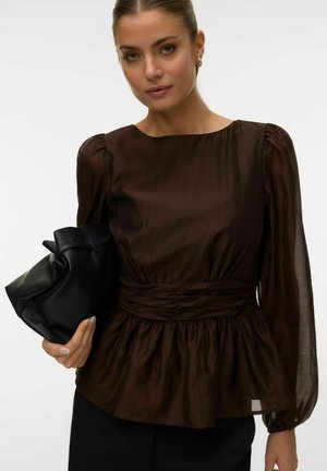 Ženska, oblečena v prosojno rjavo bluzo z dolgimi rokavi in naborkom na pasu, drži črno mehko torbico na enobarvnem ozadju.