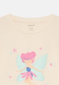Hellbeiges T-Shirt mit einer bunten Feen-Grafik mit pinken Haaren, türkisfarbenen Flügeln und floralen Akzenten. Weiches Material, Rundhalsausschnitt.