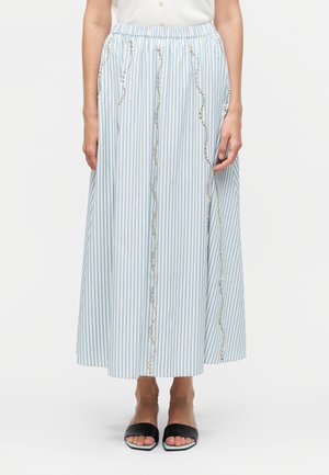 Žena nosiaca modro-bielu midi sukňu s vertikálnymi pruhmi a vlnitými ozdobami z kryštálikov, kombinovanú s čiernymi sandálmi s otvorenou špičkou.