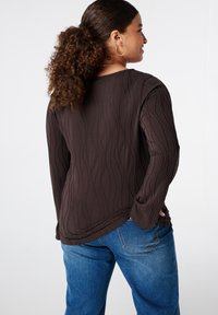 Vrouw met krullend haar in een paardenstaart draagt een donkerbruine textuur lange mouwen shirt en blauwe jeans, kijkt naar rechts met haar hand in haar zak.