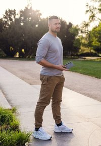 Man standing on a path, wearing a gray long-sleeve shirt, khaki pants, and white athletic shoes. He holds a gray device in one hand.