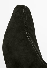 Scarpa in suede nero con punta appuntita e cuciture bianche visibili che corrono lungo il centro.