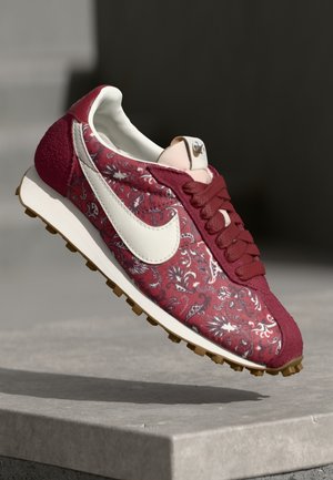 Rød og hvid Nike-sneaker med blomstermønster, hvid swoosh, bordeaux snørebånd og gummisål, vist svævende over betonoverflade.