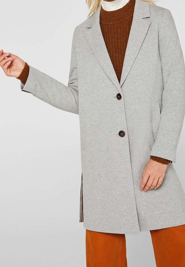 Mujer vestida con un abrigo gris claro de botones sobre un suéter marrón y un cuello de tortuga blanco, combinado con pantalones naranjas, de pie con un brazo ligeramente levantado.