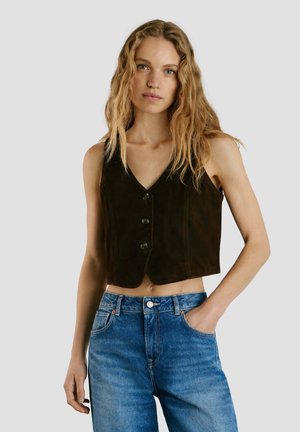 Mujer con cabello largo y ondulado rubio, vestida con un chaleco negro sin mangas abotonado y vaqueros azules de talle alto, de pie con una mano en el bolsillo.