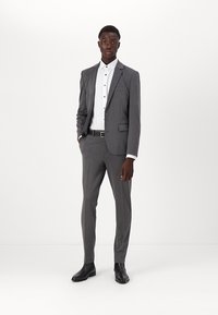Costume gris en tissu texturé, design ajusté avec une veste à un bouton, chemise blanche, boutons noirs et chaussures de ville noires.
